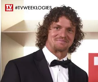 Man in a tuxedo smiling at a TV event backdrop with "TV WEEK LOGIES" hashtag in top corner.