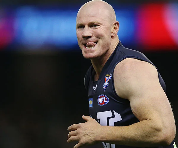 AFL player in a navy jersey sticking out his tongue on the field, with AFL and team's logos visible.