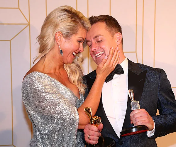 A couple joyfully embracing, holding awards, with one person touching the other's face lovingly.