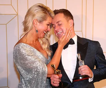 A couple joyfully embracing, holding awards, with one person touching the other's face lovingly.