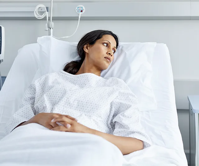 A woman in a hospital bed wearing a gown, looking contemplative.