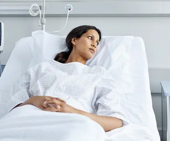 A woman in a hospital bed wearing a gown, looking contemplative.