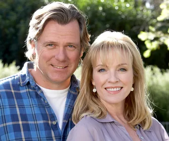 A man in a blue plaid shirt and a woman in a gray blouse smile outdoors, with green foliage in the background.