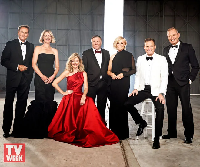 Group of seven people in formal attire posing for a professional photo, some standing and one sitting in a red dress.