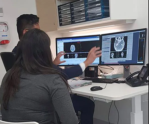 Doctors reviewing brain scan images on dual computer monitors in a medical office setting.