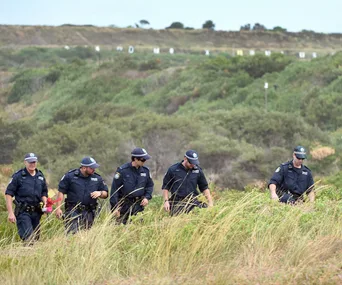 Police officers walking through grassy terrain during a search operation.