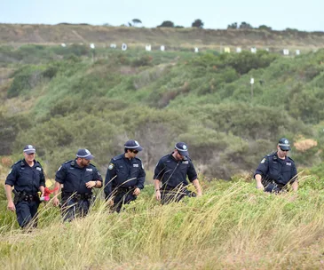 Police officers walking through grassy terrain during a search operation.