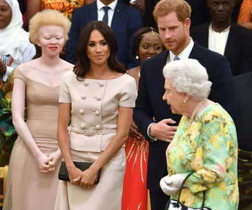 Prince Harry and Meghan Markle hold hands at an event, with attendees in the background.