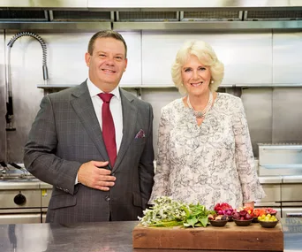 Two people in a kitchen standing behind a cutting board with herbs and vegetables.
