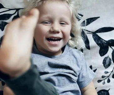 Smiling child with blond hair and a gray shirt, playfully raising a foot, surrounded by floral-patterned cushions.