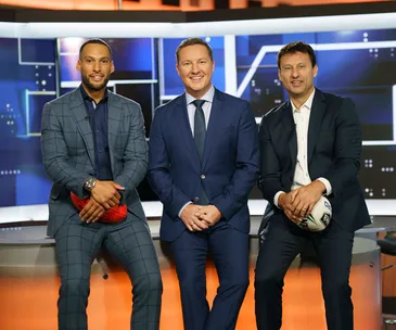 Three men in suits sitting on a news set desk, each holding a sports ball.