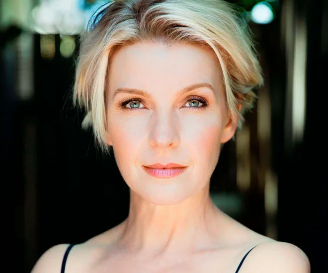 A woman with short blonde hair and a neutral expression, set against a blurred outdoor background.
