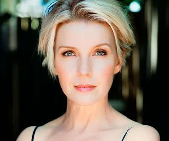 A woman with short blonde hair and a neutral expression, set against a blurred outdoor background.