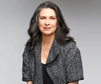 Pamela Rabe, with long hair, in a gray tweed jacket, on a neutral background.