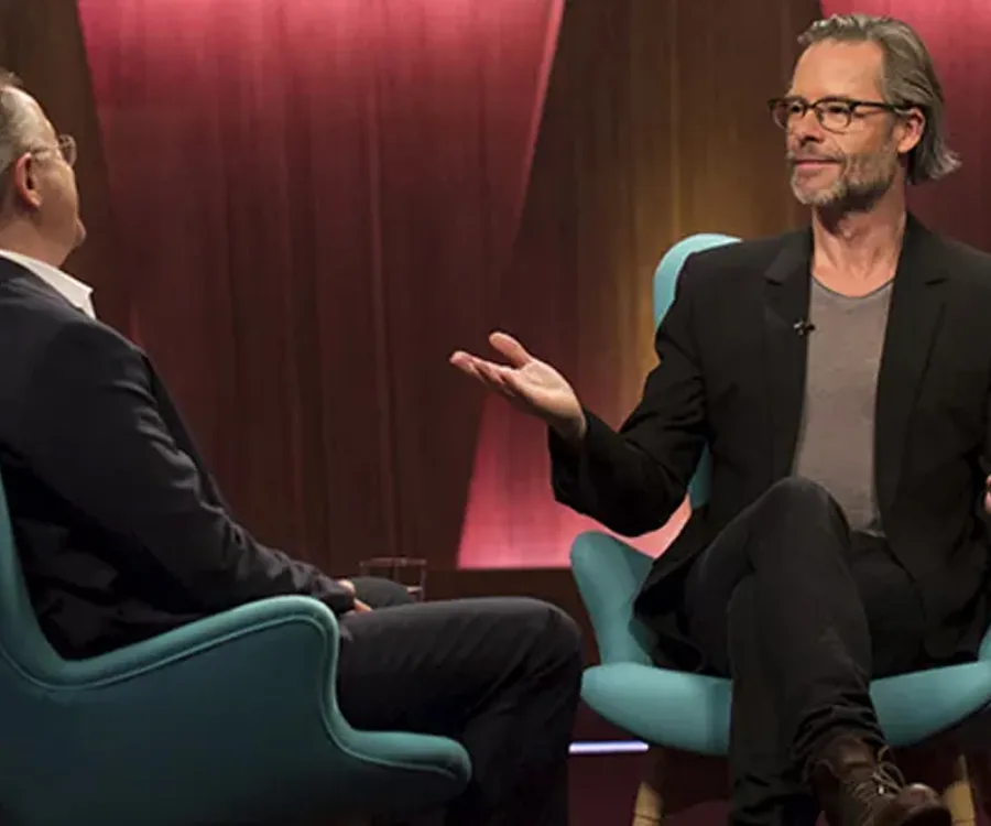 Guy Pearce in conversation on a talk show set, seated casually with a host opposite.