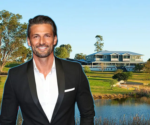 Man in a suit standing in front of a large house with a pond and trees in the background.