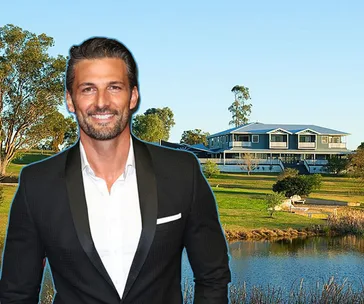 Man in a suit standing in front of a large house with a pond and trees in the background.