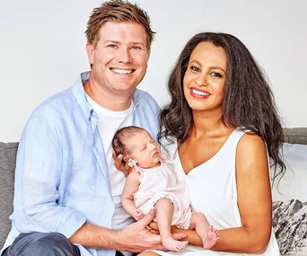 Couple poses with their newborn baby, both smiling warmly, creating a family portrait on a neutral background.