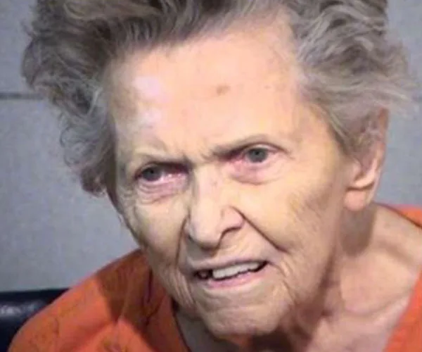 An elderly woman with short hair and wearing an orange top, appearing distressed in a mugshot.