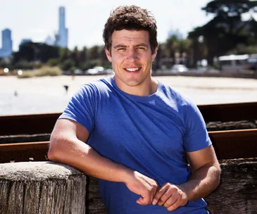 A person in a blue shirt smiles while leaning against a wooden beam at a beach, with a city skyline in the background.