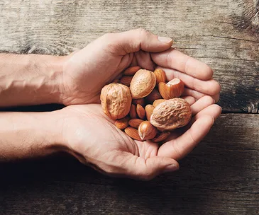 Hands holding various nuts, including almonds and walnuts, over a wooden surface.