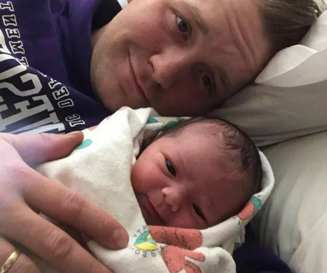 Father cuddling newborn daughter wrapped in a blanket, both smiling gently at the camera while lying on a bed.