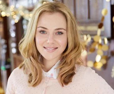 Blonde woman smiling in a softly lit indoor setting with bokeh lights in the background.