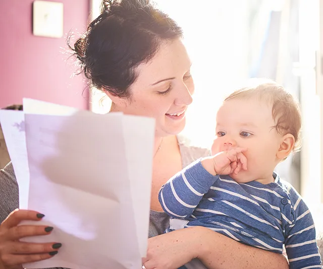 A smiling woman holds a baby while reading a document, with sunlight in the background.