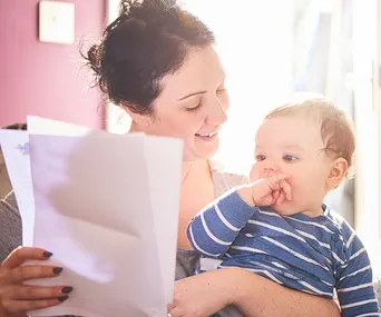 A smiling woman holds a baby while reading a document, with sunlight in the background.