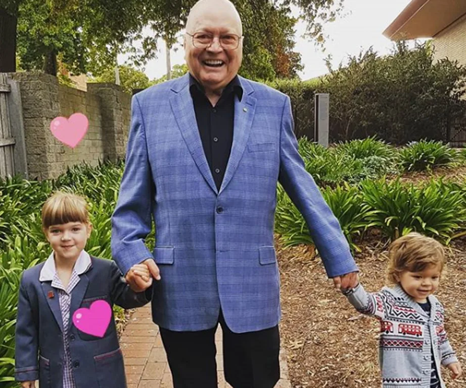 Man in blue blazer holding hands with two young children on a garden path, pink heart nearby.