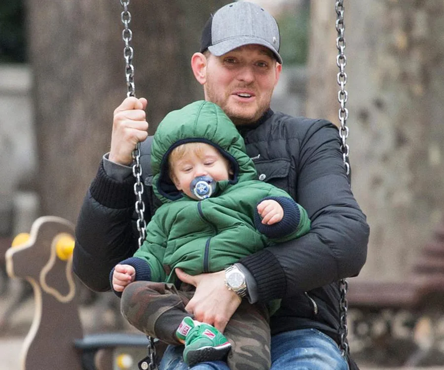 A man and a child on a swing in a park; the child is wearing a green jacket and pacifier.