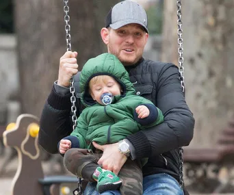 A man and a child on a swing in a park; the child is wearing a green jacket and pacifier.
