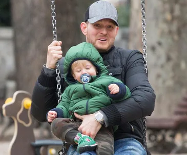 A man and a child on a swing in a park; the child is wearing a green jacket and pacifier.