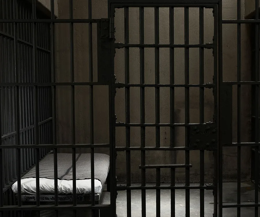 A dimly lit prison cell with a metal bed frame, narrow mattress, and barred door.
