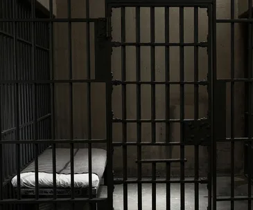 A dimly lit prison cell with a metal bed frame, narrow mattress, and barred door.