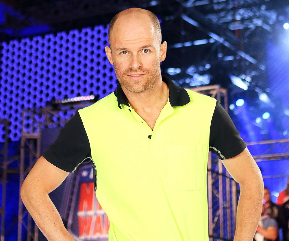 Man in high-visibility shirt at Australian Ninja Warrior course, standing confidently against a blue-lit obstacle backdrop.