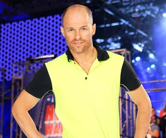Man in high-visibility shirt at Australian Ninja Warrior course, standing confidently against a blue-lit obstacle backdrop.
