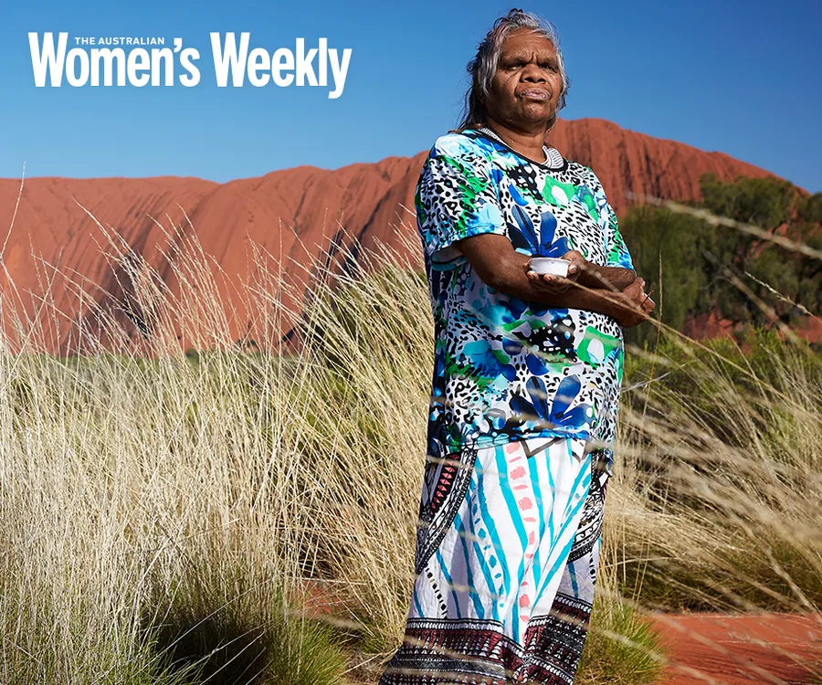 Indigenous artist stands in colorful dress holding an object, with Uluru in the background, under clear blue sky.