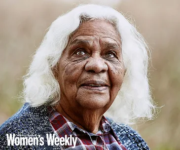 Elderly woman with white hair and gentle expression, wearing a blue sweater over a plaid shirt, featured in Women’s Weekly.