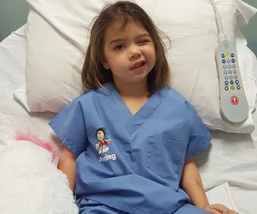 A young child in a hospital bed, wearing blue scrubs, with a hospital remote nearby.