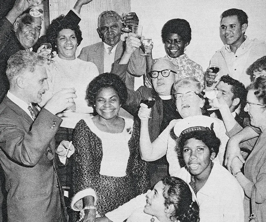 Group of people in a celebratory setting, raising glasses and smiling, black and white photo from the 1960s.
