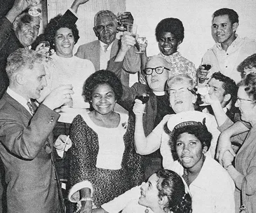 Group of people in a celebratory setting, raising glasses and smiling, black and white photo from the 1960s.