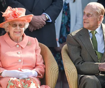 Two elderly people seated outdoors, one in a peach outfit and hat, the other in a suit, smiling at each other.