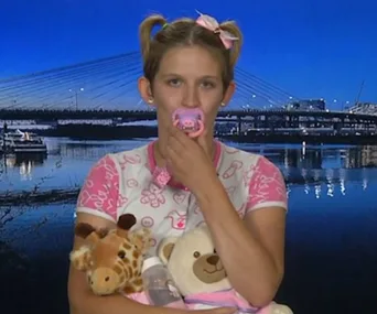 Young woman with pacifier, hair in pigtails, holding stuffed giraffe and teddy bear, standing in front of a lit bridge backdrop.