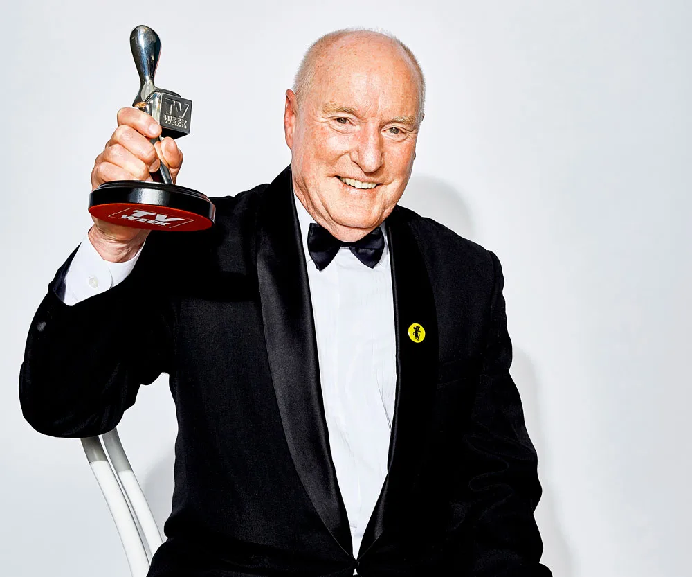 A man in a tuxedo smiles while holding an award trophy, with a white background.