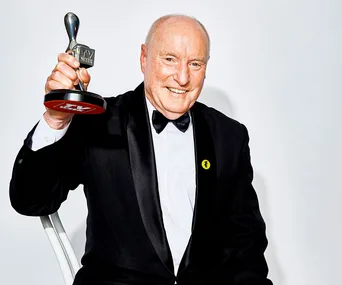 A man in a tuxedo smiles while holding an award trophy, with a white background.