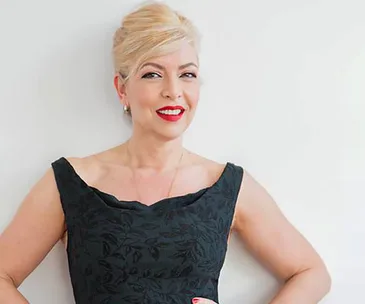 A woman with blonde hair and red lipstick in a black dress stands smiling against a plain background.