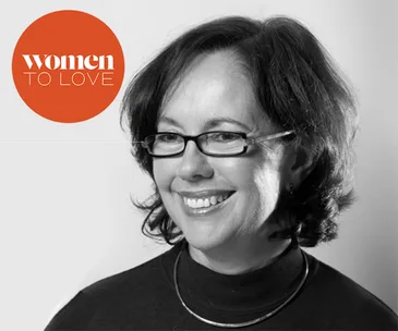 A woman smiles in a black and white portrait, wearing glasses and a necklace, with "Women to Love" logo in the top left corner.
