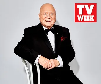 Elderly man in a tuxedo, smiling, seated on a chair with TV WEEK logo in background.