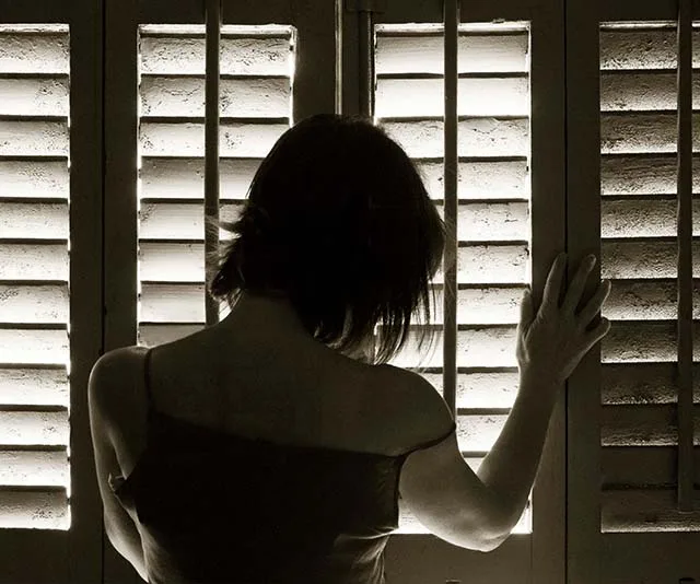 A woman stands pensively at window blinds, her hand touching the slats, silhouetted by light filtering through.